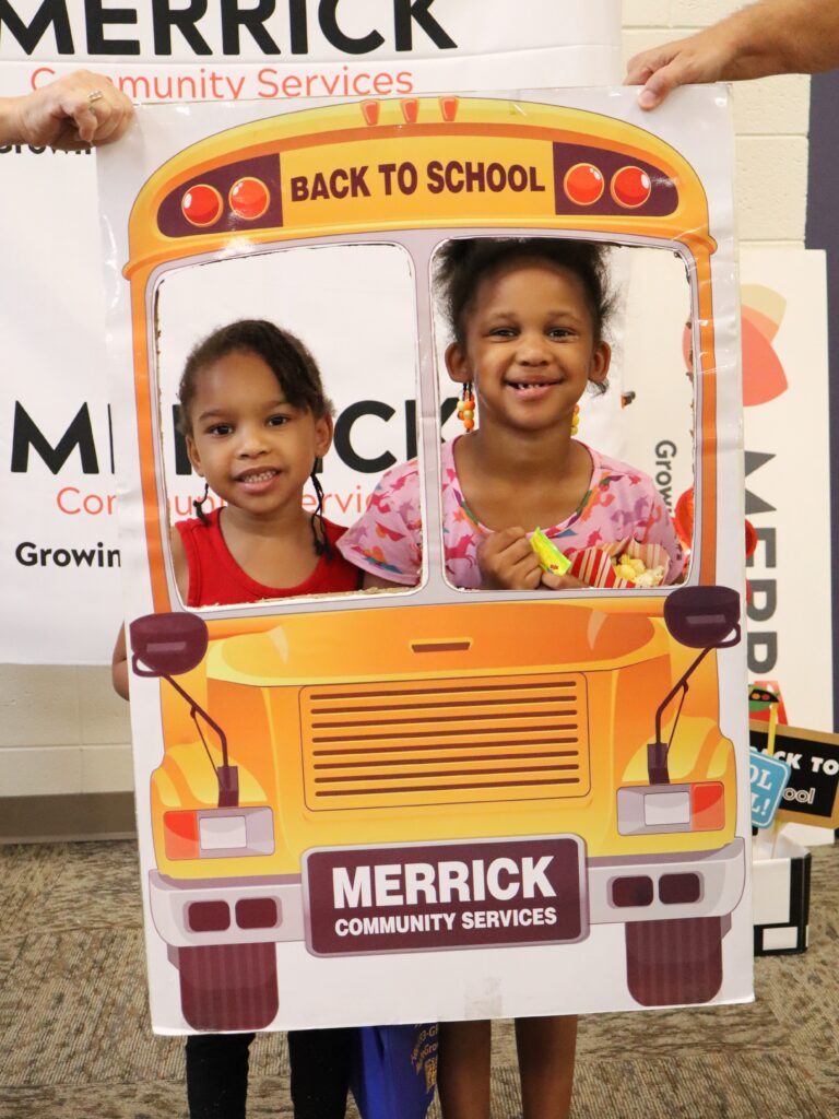 Two children smiling in a school-bus photo frame at Merrick’s Backpack Distribution
